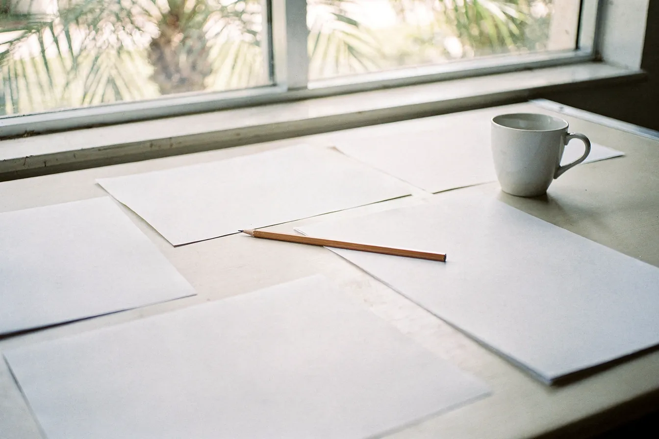 A bright drafting table with blank sheets, a T-square and a coffee cup by a window with palms