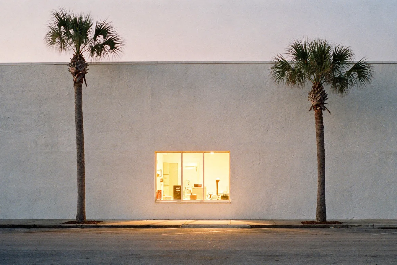 A single bright window lit in a quiet street of low white buildings at dusk