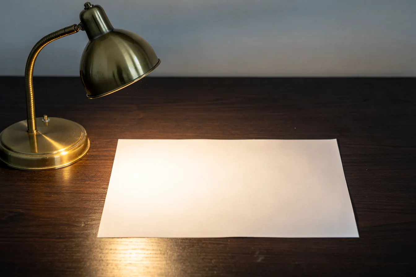 A brass desk lamp lighting a single blank sheet of paper on a dark wooden desk