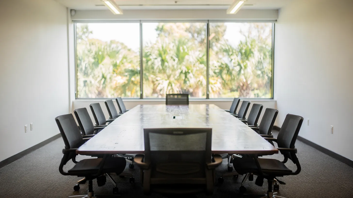 An empty conference room with a long table and black chairs in front of tall windows onto palms