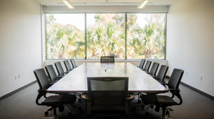 An empty conference room with a long table and black chairs in front of tall windows onto palms