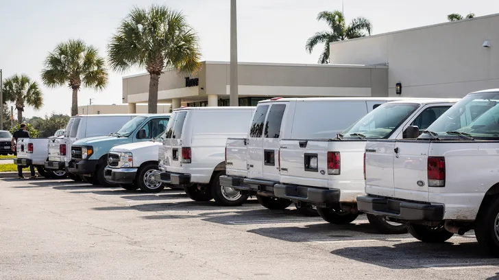 A small business parking lot in bright morning sun, most spaces filled with plain white work vans and pickup trucks, only one or two people visible in the distance.