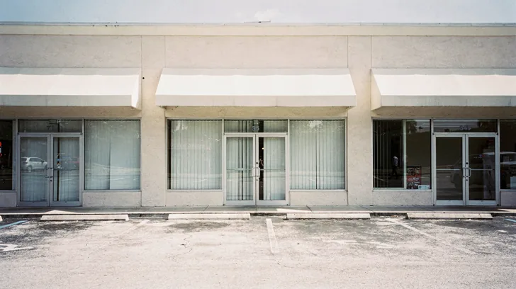 A row of Southwest Florida strip mall storefronts in flat bright midday sun, the units on either side open while the middle one sits dark with its blinds drawn.