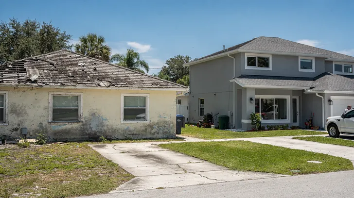 An older single-story home with a worn, stained roof beside a newly built two-story house