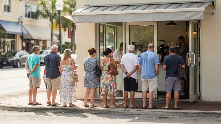 Customers lined up along the sidewalk outside a small cafe with its front doors open, seen from behind