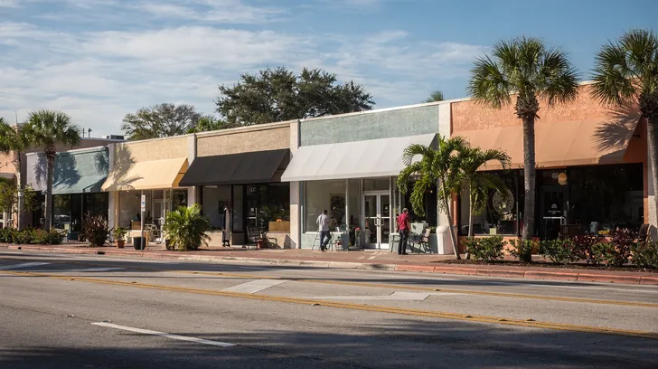 A row of small storefronts with plain awnings on a sunny boulevard lined with palms, seen from across the street