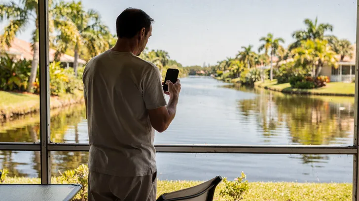 A homeowner on a screened lanai holding up a phone, seen from behind, a canal and palms beyond