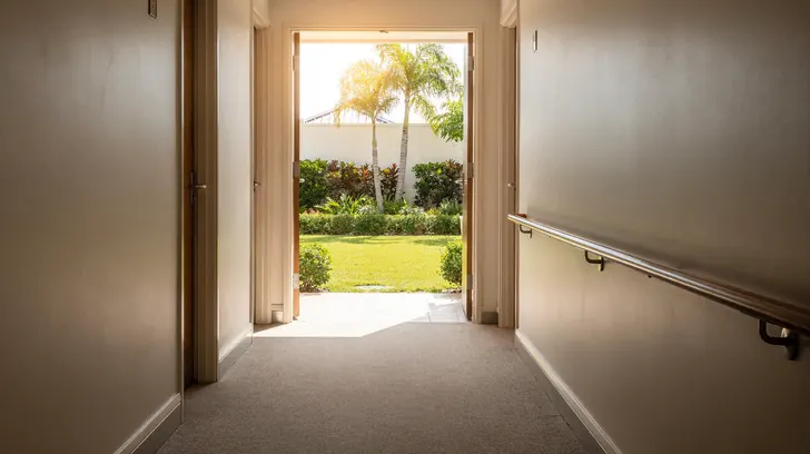 A quiet sunlit corridor in a single storey Florida senior living community, a wooden handrail along one wall and an open doorway onto a garden courtyard with royal palms.