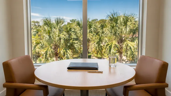 A bright private meeting room in a Naples office, a round wooden table with two leather chairs and a closed leather folio, a tall window behind looking out onto palm trees against a blue sky.