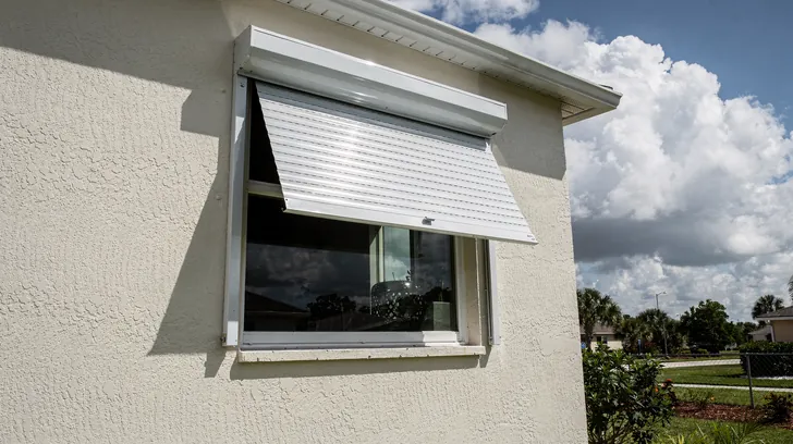 A storm shutter half lowered over the window of a stucco home as clouds build in the summer sky