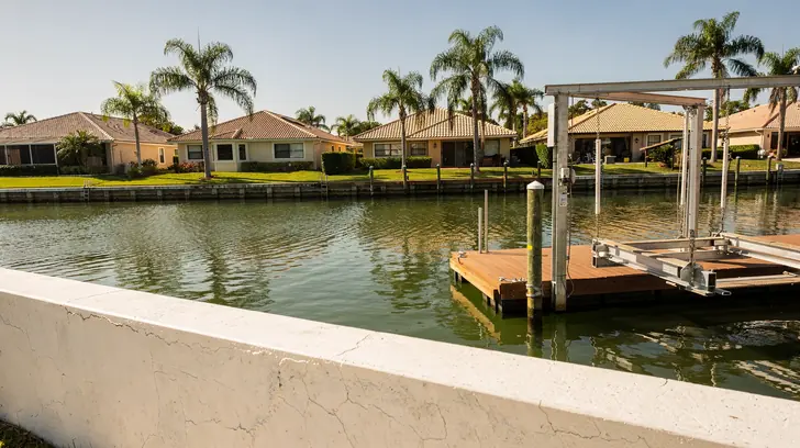 A concrete seawall cap and a timber dock on a Southwest Florida residential canal in bright morning sun, a boat lift beside the dock and single storey homes along the far bank.