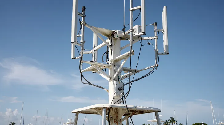A white mast of radio antennas and receivers against a bright blue sky above a marina
