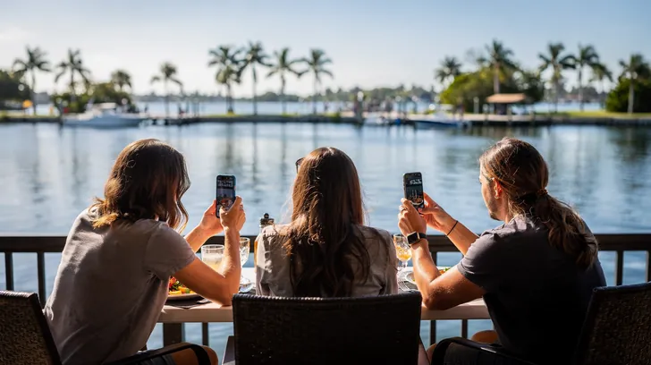 Three friends at a waterfront table photographing their food with phones, seen from behind, palms across the water