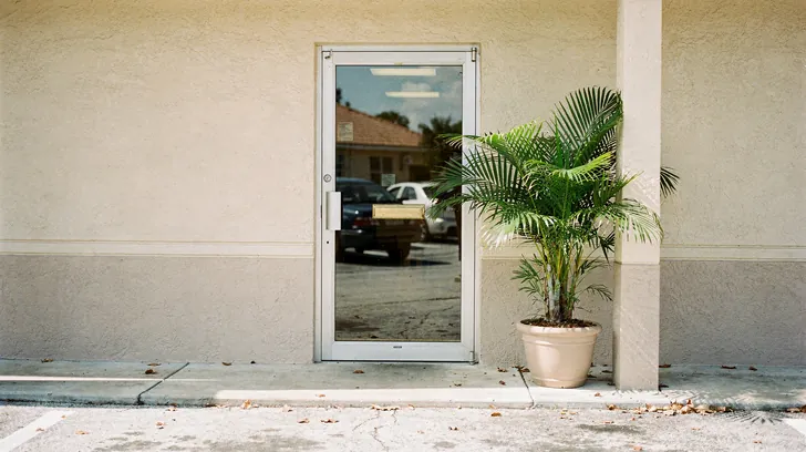 The plain glass door of a small stucco office with a potted palm beside it, parked cars reflected in the glass